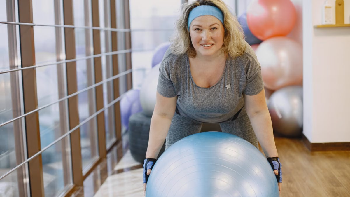 An adult woman engaging in a fitness workout with an exercise ball in a gym setting.